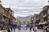 A bustling street with traditional buildings on either side, adorned with colorful prayer flags strung across. People walk along the street, some carrying bags, while others browse shopfronts displaying various goods. A prominent sign in the foreground reads 'SMILE YOU ARE UNDER CCTV'. In the background, there are snow-capped mountains under a cloudy sky.