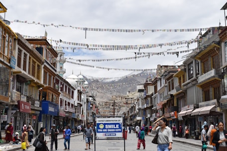 A bustling street with traditional buildings on either side, adorned with colorful prayer flags strung across. People walk along the street, some carrying bags, while others browse shopfronts displaying various goods. A prominent sign in the foreground reads 'SMILE YOU ARE UNDER CCTV'. In the background, there are snow-capped mountains under a cloudy sky.