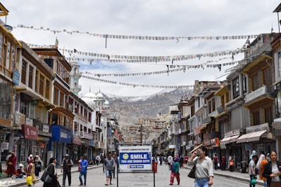A bustling street with traditional buildings on either side, adorned with colorful prayer flags strung across. People walk along the street, some carrying bags, while others browse shopfronts displaying various goods. A prominent sign in the foreground reads 'SMILE YOU ARE UNDER CCTV'. In the background, there are snow-capped mountains under a cloudy sky.