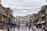 A bustling street with traditional buildings on either side, adorned with colorful prayer flags strung across. People walk along the street, some carrying bags, while others browse shopfronts displaying various goods. A prominent sign in the foreground reads 'SMILE YOU ARE UNDER CCTV'. In the background, there are snow-capped mountains under a cloudy sky.