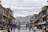 A bustling street with traditional buildings on either side, adorned with colorful prayer flags strung across. People walk along the street, some carrying bags, while others browse shopfronts displaying various goods. A prominent sign in the foreground reads 'SMILE YOU ARE UNDER CCTV'. In the background, there are snow-capped mountains under a cloudy sky.