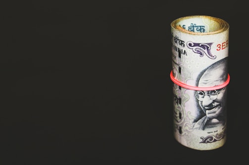 A rolled-up bundle of Indian currency notes secured with a red rubber band is placed against a solid, dark background. The banknotes prominently feature a portrait of Mahatma Gandhi.