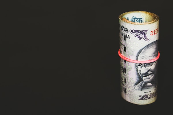 A rolled-up bundle of Indian currency notes secured with a red rubber band is placed against a solid, dark background. The banknotes prominently feature a portrait of Mahatma Gandhi.