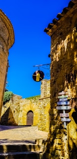 A quaint, narrow street lined with ancient stone buildings under a clear blue sky. The architecture reflects a historical style with rustic stonework. A hanging metal sign features what appears to be a bell. A multi-directional signpost displays various words in French and English such as 'cadeaux', 'gifts', and 'bijoux'. Sunlight casts dynamic shadows on the textured walls.