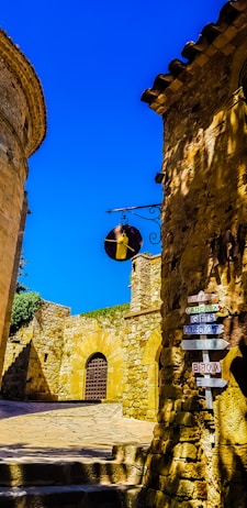 A quaint, narrow street lined with ancient stone buildings under a clear blue sky. The architecture reflects a historical style with rustic stonework. A hanging metal sign features what appears to be a bell. A multi-directional signpost displays various words in French and English such as 'cadeaux', 'gifts', and 'bijoux'. Sunlight casts dynamic shadows on the textured walls.