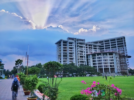 A large modern building under construction is situated near a green field. There are people walking on a paved path and others playing or walking on the grass. The sky above is partly cloudy with sun rays peeking through the clouds, creating a dramatic effect. There are small trees and vibrant pink flowers in the foreground.