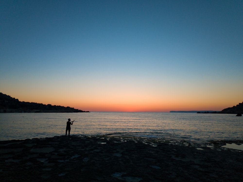person standing near seashore viewing blue sea under blue and orange skies
