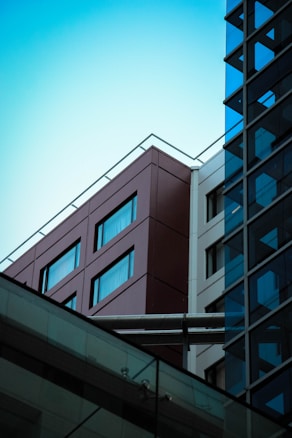 Two modern buildings intersect, one with a sleek glass exterior reflecting the light and sky, and the other with a brown facade featuring rectangular windows. A bridging structure connects the buildings, and the bright sky creates a contrasting backdrop.