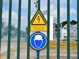 A fenced area with warning signs is visible. The yellow triangular sign includes a lightning bolt symbol and the German text 'Hochspannung Lebensgefahr,' indicating high voltage and danger to life. Below it is a blue circular sign with an icon of a person wearing a hard hat, suggesting mandatory safety equipment. Behind the fence, there are electrical installations, possibly part of a transformer station.