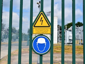 A fenced area with warning signs is visible. The yellow triangular sign includes a lightning bolt symbol and the German text 'Hochspannung Lebensgefahr,' indicating high voltage and danger to life. Below it is a blue circular sign with an icon of a person wearing a hard hat, suggesting mandatory safety equipment. Behind the fence, there are electrical installations, possibly part of a transformer station.