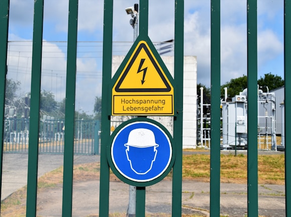 A fenced area with warning signs is visible. The yellow triangular sign includes a lightning bolt symbol and the German text 'Hochspannung Lebensgefahr,' indicating high voltage and danger to life. Below it is a blue circular sign with an icon of a person wearing a hard hat, suggesting mandatory safety equipment. Behind the fence, there are electrical installations, possibly part of a transformer station.
