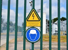 A fenced area with warning signs is visible. The yellow triangular sign includes a lightning bolt symbol and the German text 'Hochspannung Lebensgefahr,' indicating high voltage and danger to life. Below it is a blue circular sign with an icon of a person wearing a hard hat, suggesting mandatory safety equipment. Behind the fence, there are electrical installations, possibly part of a transformer station.