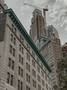 Tall skyscrapers under construction are captured from a low angle, with a visible crane towering above the buildings. Architectural details of older buildings in the foreground contrast with the modern structures in the background. The sky is overcast, creating a moody atmosphere.