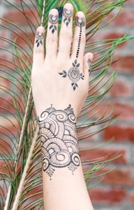 A hand adorned with intricate black henna designs, featuring floral and geometric patterns, resting against a peacock feather with a blurred brick wall in the background.