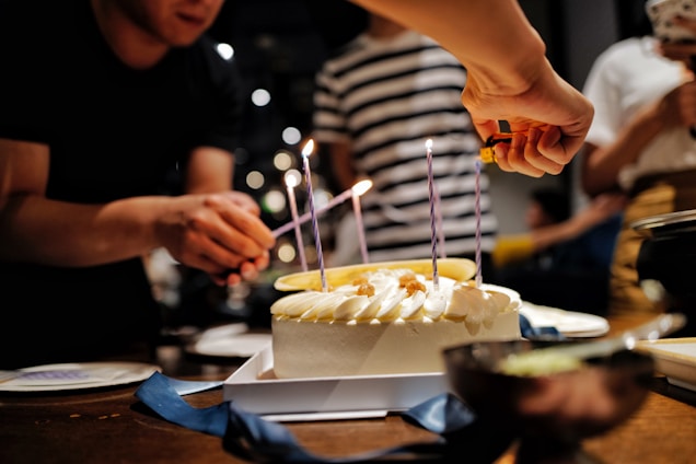 A birthday cake with white frosting and purple candles is being lit by a hand holding a lighter. People are gathered around the table, partially visible, with one person holding a match to light the candles. The setting appears festive and informal.