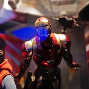 A close-up of a detailed Iron Man action figure, with its iconic red and gold armor. The figure is illuminated with dramatic lighting, highlighting its metallic texture and features. To the left, part of a person wearing a red hoodie is visible, suggesting the figure is on display or being observed. The background is blurred, with hints of blue and red lighting effects, enhancing the futuristic theme.