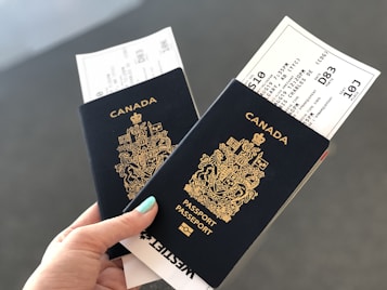 A hand holding two Canadian passports, each containing a boarding pass. The passports are navy blue with gold text and the Canadian coat of arms embossed on the cover. The background is neutral and out of focus.