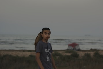 A young person stands near a beach with the ocean in the background. They are wearing a t-shirt that has text on it and have a straw hat slung over their shoulder. The sand and sparse vegetation are visible in the foreground, with a small tent pitched in the distance.