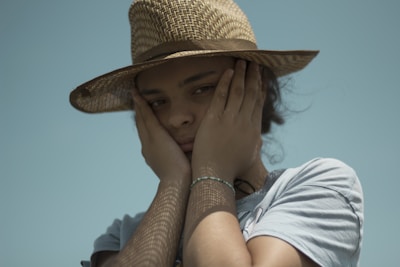 A person wearing a light gray shirt and a woven straw hat covers their cheeks with both hands, exuding a contemplative or melancholic expression. The background is a clear, pale blue sky, and the lighting creates a play of shadows on the face and hat.
