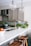 A modern kitchen with beige cabinets, a white marble countertop, and brass fixtures. There are two wooden stools at the counter, a bowl of apples, and various green vegetables and herbs placed near the sink. Two large glass pendant lights hang from the ceiling.