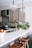 A modern kitchen with beige cabinets, a white marble countertop, and brass fixtures. There are two wooden stools at the counter, a bowl of apples, and various green vegetables and herbs placed near the sink. Two large glass pendant lights hang from the ceiling.