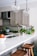 A modern kitchen with beige cabinets, a white marble countertop, and brass fixtures. There are two wooden stools at the counter, a bowl of apples, and various green vegetables and herbs placed near the sink. Two large glass pendant lights hang from the ceiling.