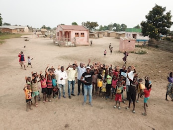 A group of people stands on a sandy open area in a residential setting. There are several children and adults, all looking up and waving toward the camera. A few houses with pink and beige walls are in the background, surrounded by small patches of greenery. The setting appears to be a rural village, with simple structures and a dirt field.