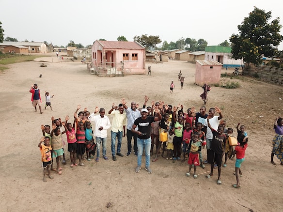 A group of people stands on a sandy open area in a residential setting. There are several children and adults, all looking up and waving toward the camera. A few houses with pink and beige walls are in the background, surrounded by small patches of greenery. The setting appears to be a rural village, with simple structures and a dirt field.