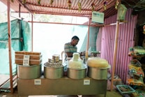 A small street stall with various traditional sweets and snacks displayed in large metal containers. A man in casual attire stands behind the counter, partially obscured by the items. Signs indicating digital payment options like Paytm are visible. The stall is housed within a simple structure made of corrugated metal and cloth.