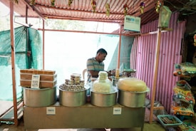 A small street stall with various traditional sweets and snacks displayed in large metal containers. A man in casual attire stands behind the counter, partially obscured by the items. Signs indicating digital payment options like Paytm are visible. The stall is housed within a simple structure made of corrugated metal and cloth.