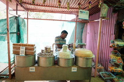 A small street stall with various traditional sweets and snacks displayed in large metal containers. A man in casual attire stands behind the counter, partially obscured by the items. Signs indicating digital payment options like Paytm are visible. The stall is housed within a simple structure made of corrugated metal and cloth.