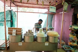 A small street stall with various traditional sweets and snacks displayed in large metal containers. A man in casual attire stands behind the counter, partially obscured by the items. Signs indicating digital payment options like Paytm are visible. The stall is housed within a simple structure made of corrugated metal and cloth.
