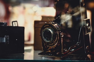 A vintage camera is prominently displayed, its intricate details and metallic components highlighted. The background is softly blurred, giving a nostalgic and classic feel to the setting.
