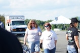 A group of people walking in a parking lot with a U-Haul truck and a white tent in the background. Two individuals are prominently featured in the foreground, one with long blond hair wearing sunglasses and a shirt that reads 'Rose Lavelle is good at soccer,' while the other person has short hair and is wearing reflective sunglasses and a different shirt. Another person to the right has a cap and is wearing a black shirt.