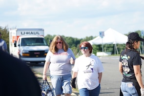 A group of people walking in a parking lot with a U-Haul truck and a white tent in the background. Two individuals are prominently featured in the foreground, one with long blond hair wearing sunglasses and a shirt that reads 'Rose Lavelle is good at soccer,' while the other person has short hair and is wearing reflective sunglasses and a different shirt. Another person to the right has a cap and is wearing a black shirt.
