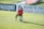 A soccer player is kneeling on a grassy field holding a soccer ball, wearing a green and red uniform with gloves. There are advertising banners in the background, along with a training center sign.
