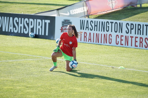 A soccer player is kneeling on a grassy field holding a soccer ball, wearing a green and red uniform with gloves. There are advertising banners in the background, along with a training center sign.