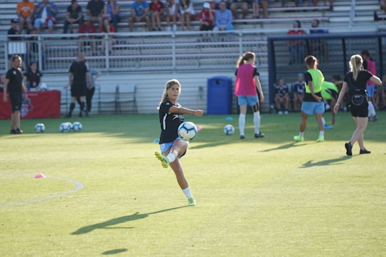 A group of female soccer players is practicing on a grass field, with one player in the foreground performing a dynamic kick with a soccer ball mid-air. Other players and equipment can be seen in the background, along with spectators sitting on bleachers.