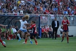 A soccer match is taking place with players from opposing teams engaged in action on the field. The player in white is attempting a maneuver with the ball while an opposing player in black and red tries to intercept. The crowd in the background is attentive, and team members are seated on the sidelines. A coach or official stands by observing the game.