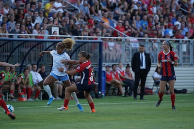 A soccer match is taking place with players from opposing teams engaged in action on the field. The player in white is attempting a maneuver with the ball while an opposing player in black and red tries to intercept. The crowd in the background is attentive, and team members are seated on the sidelines. A coach or official stands by observing the game.