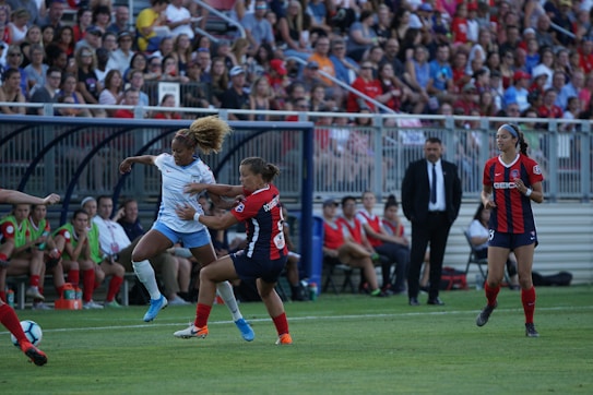 A soccer match is taking place with players from opposing teams engaged in action on the field. The player in white is attempting a maneuver with the ball while an opposing player in black and red tries to intercept. The crowd in the background is attentive, and team members are seated on the sidelines. A coach or official stands by observing the game.
