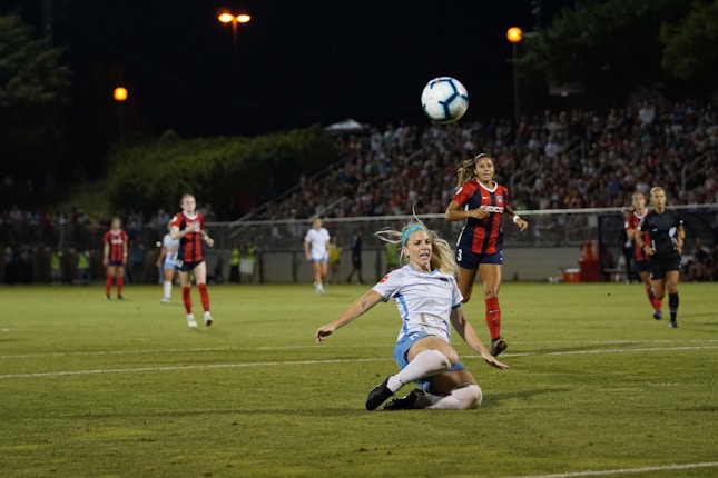 A soccer match at night with one player in a white and blue uniform executing a slide tackle while others in red and blue uniforms watch. The crowd in the background and stadium lights create an intense atmosphere.