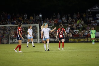 A diverse group of women soccer players celebrating together on a lush green field under bright stadium lights.