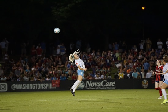 A soccer match scene with a player in mid-air attempting a header. The background shows a crowd of spectators. The setting is a well-lit field during nighttime, with players wearing uniforms in different colors.