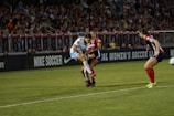 A soccer match is taking place with players in action, one wearing a white and light blue uniform and two others in red and blue uniforms. They are surrounded by a crowded stadium with bright lights and advertisements, indicating possible sponsorships like Nike.
