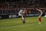 A soccer match is taking place with players in action, one wearing a white and light blue uniform and two others in red and blue uniforms. They are surrounded by a crowded stadium with bright lights and advertisements, indicating possible sponsorships like Nike.