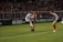 A soccer match is taking place with players in action, one wearing a white and light blue uniform and two others in red and blue uniforms. They are surrounded by a crowded stadium with bright lights and advertisements, indicating possible sponsorships like Nike.