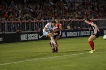 A soccer match is taking place with players in action, one wearing a white and light blue uniform and two others in red and blue uniforms. They are surrounded by a crowded stadium with bright lights and advertisements, indicating possible sponsorships like Nike.