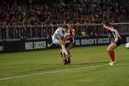 A soccer match is taking place with players in action, one wearing a white and light blue uniform and two others in red and blue uniforms. They are surrounded by a crowded stadium with bright lights and advertisements, indicating possible sponsorships like Nike.