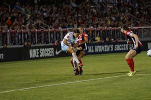 A soccer match is taking place with players in action, one wearing a white and light blue uniform and two others in red and blue uniforms. They are surrounded by a crowded stadium with bright lights and advertisements, indicating possible sponsorships like Nike.